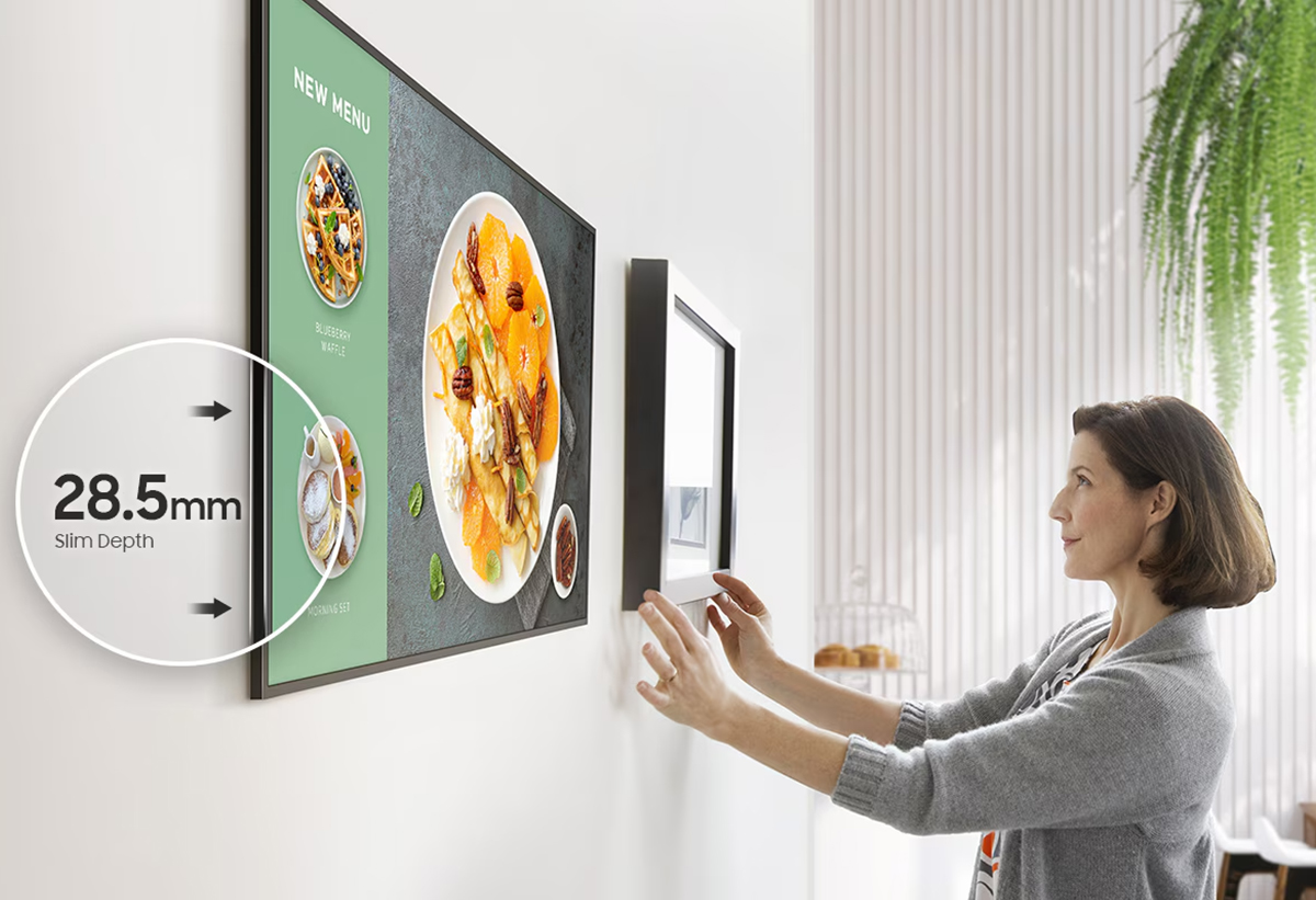 Απεικονίζεται γυναίκα να τοποθετεί την οθόνη σε τοίχο, δίπλα σε κάδρο που φαίνεται πιο φαρδύ από την οθόνη.